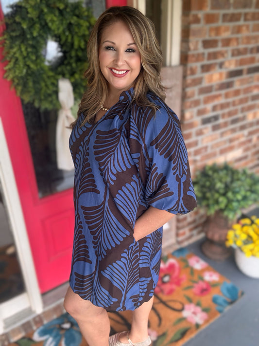 A woman modeling a short-sleeve, button-front shift dress with a blue and brown tropical leaf pattern, standing on a brick porch.