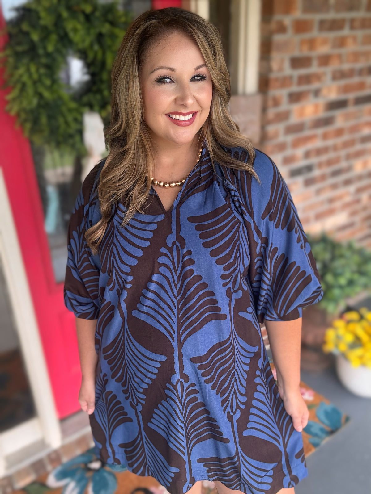 A woman wearing a short-sleeve blue and brown tropical leaf print tunic dress standing in front of a brick wall and potted plants.