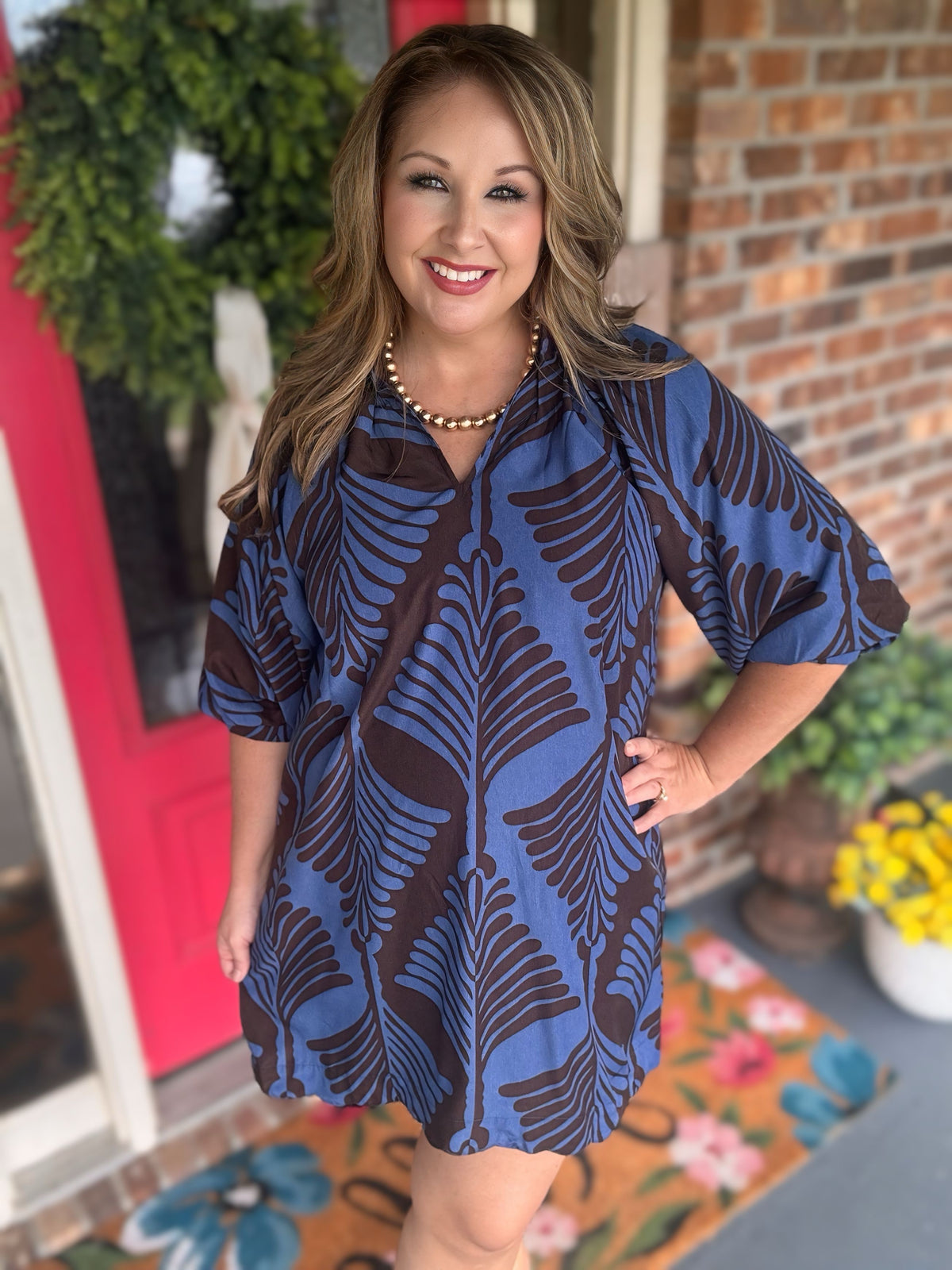 A woman modeling a short blue and brown patterned tunic dress on a front porch with a brick wall, red door, and potted plants in the background.