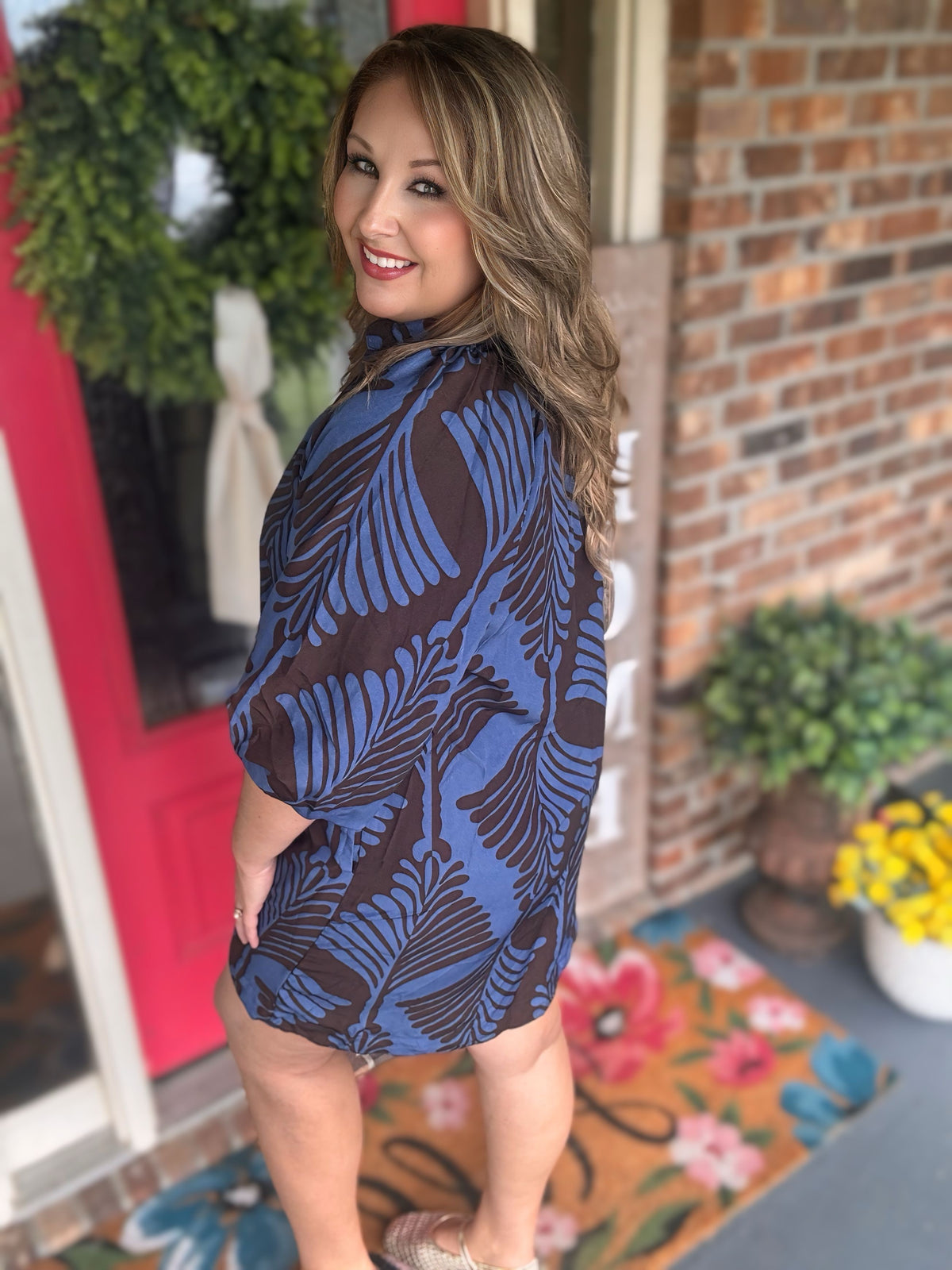 A woman wearing a short-sleeve blue and brown tropical leaf print tunic dress standing in front of a brick wall and potted plants.