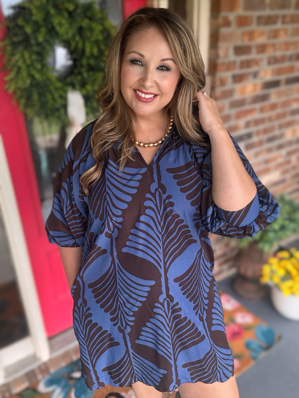 A woman wearing a short-sleeve blue and brown tropical leaf print tunic dress standing in front of a brick wall and potted plants.